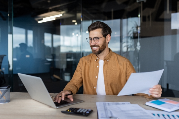 Un homme qui sourit en tapant sur son ordinateur portable et en tenant une feuille de papier.
